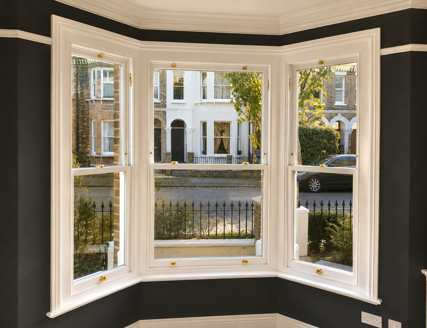 White timber bay sash windows with brass fasteners against dark charcoal walls, looking onto a railed street