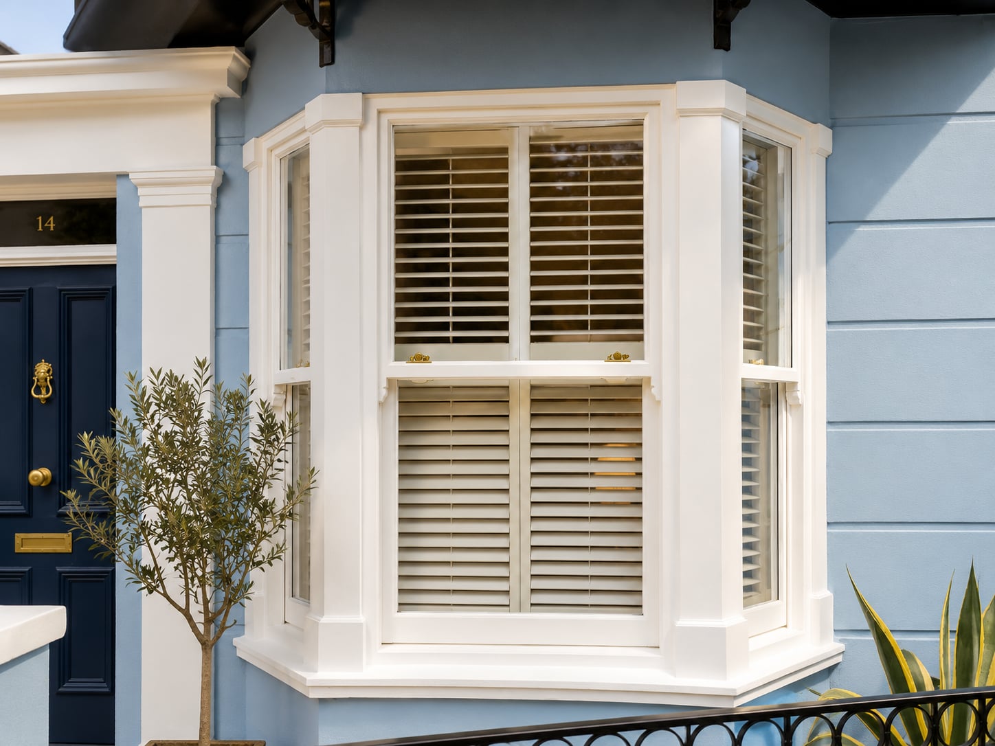 White timber bay sash window with interior shutters set into pale blue weatherboarding beside a navy door