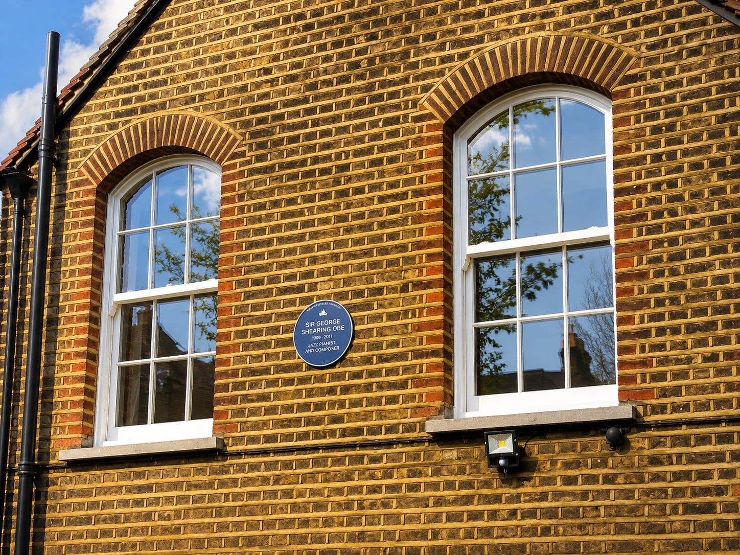 Two arched white timber sash windows under gauged brick arches on yellow stock brick, beside a blue plaque