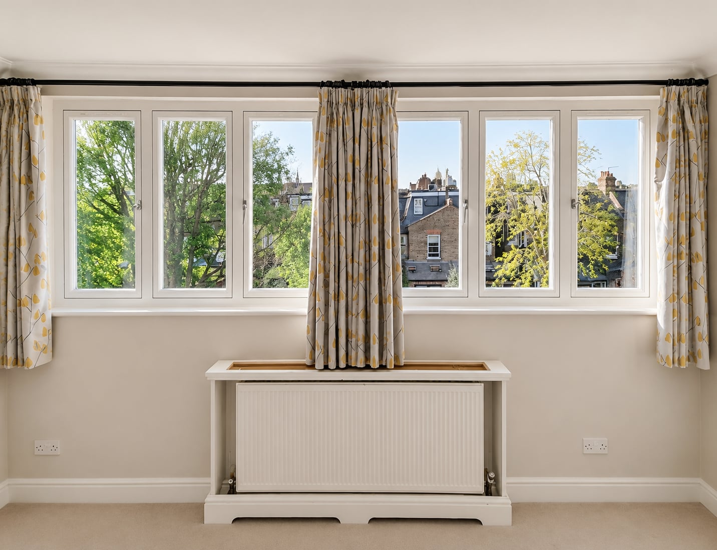 A run of white timber casement windows above a radiator seat, framing treetops and rooftops