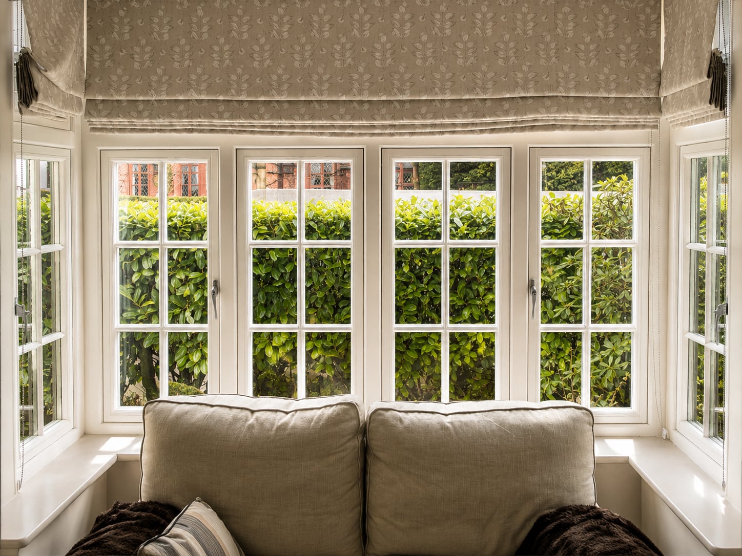 White timber bay casement windows with glazing bars above a sofa, looking onto a clipped hedge